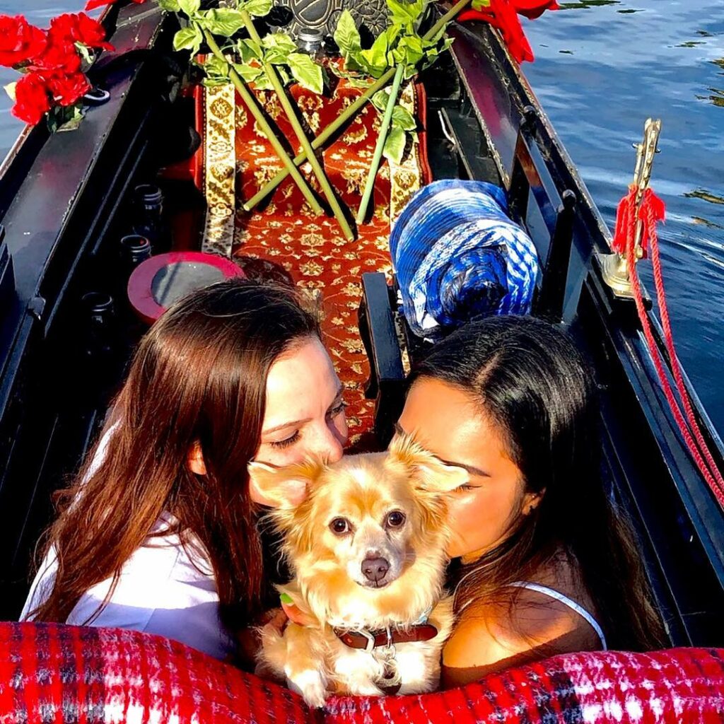 Two women sharing a romantic moment on a gondola, surrounded by rose petals and a small dog, with a scenic water backdrop, highlighting a unique date experience at Black Swan Gondola in San Diego.