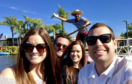 Group of friends enjoying a gondola ride on Lake San Marcos with a gondolier in a striped shirt and straw hat, surrounded by palm trees and clear blue sky, perfect for a San Diego date night experience.