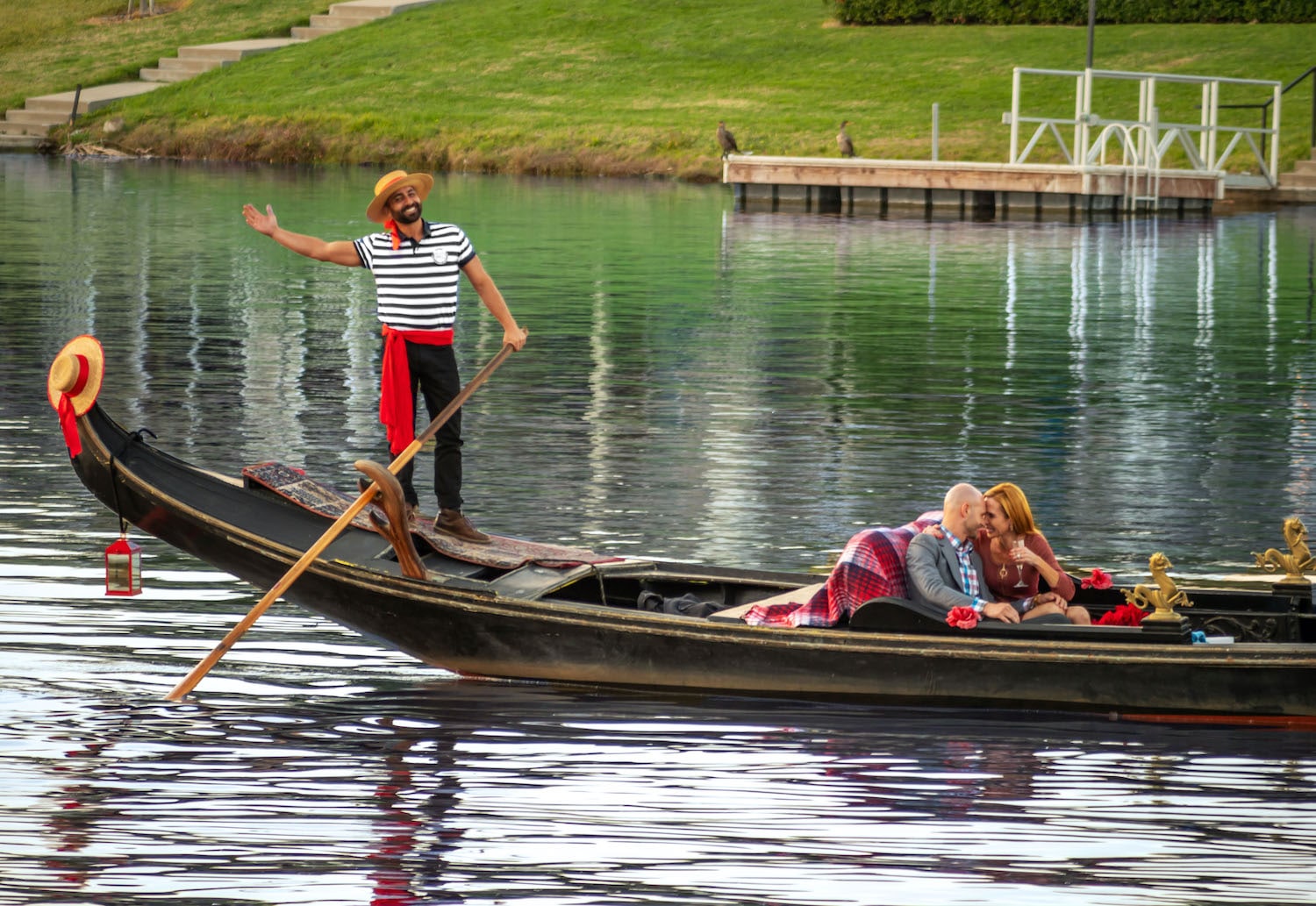 Gondola ride on serene water featuring a gondolier in striped shirt and hat, a couple enjoying a romantic moment with drinks, surrounded by lush greenery, representing anniversary celebration in San Diego.