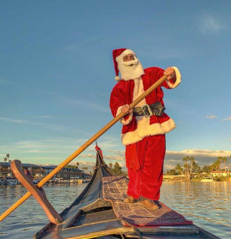 Santa Claus in a red costume rowing a gondola on a scenic lake during sunset, promoting the holiday lights cruise experience in San Diego.