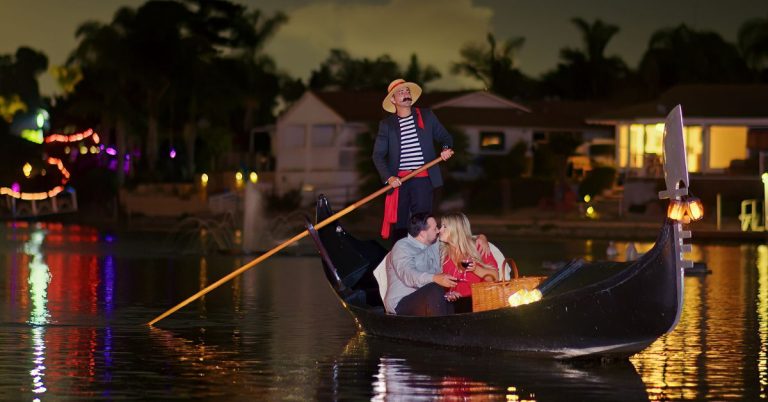 Couple enjoying a romantic gondola ride at night with a gondolier, surrounded by illuminated waterfront homes, emphasizing the intimate dining experience offered by Black Swan Gondola in San Diego.
