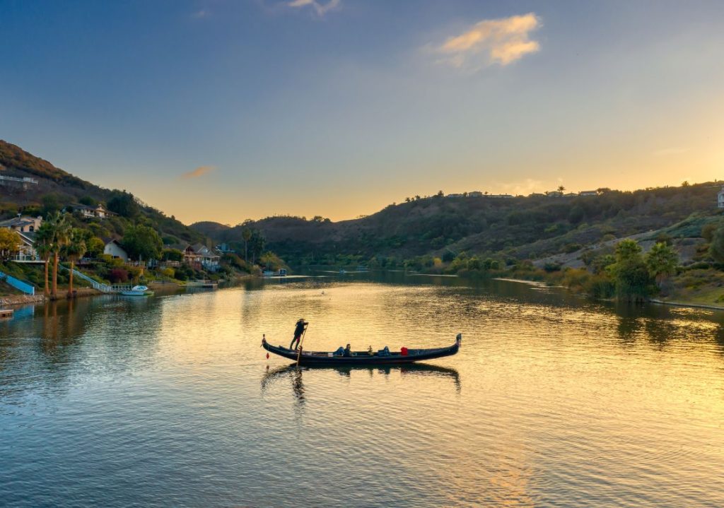 Gondola ride on Lake San Marcos at sunset with gondolier navigating tranquil waters, surrounded by lush landscapes and serene reflections, showcasing the romantic experience offered by Black Swan Gondola.