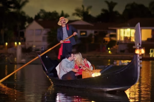 Couple sharing a romantic moment on a gondola during a San Diego dinner cruise, with a gondolier rowing in the background, surrounded by softly lit homes along the water.