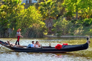 Couple enjoying a romantic gondola ride on a scenic waterway, with a gondolier guiding the boat, surrounded by lush greenery and a serene atmosphere, reflecting the unique dining experience offered by Black Swan Gondola in San Diego.